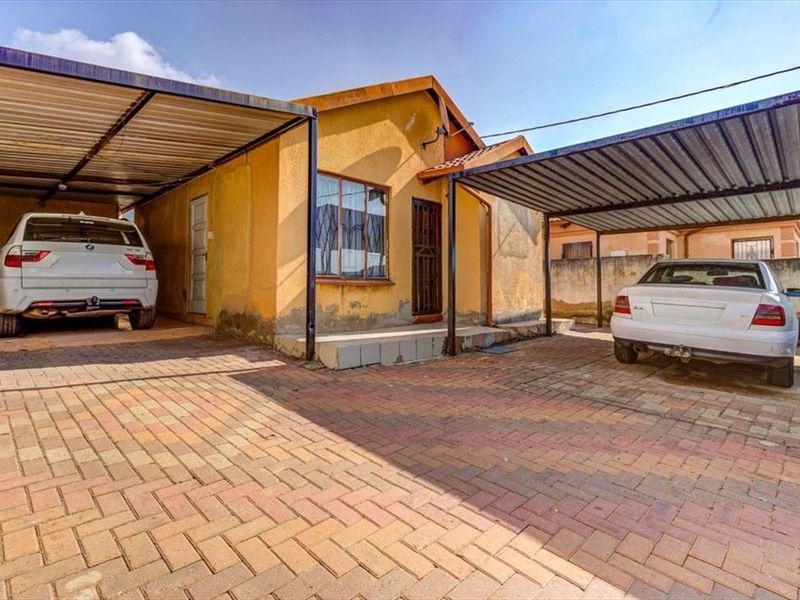 Front exterior view of the house featuring a paved driveway and two covered carports, showing signs of weathered exterior paint and plaster.