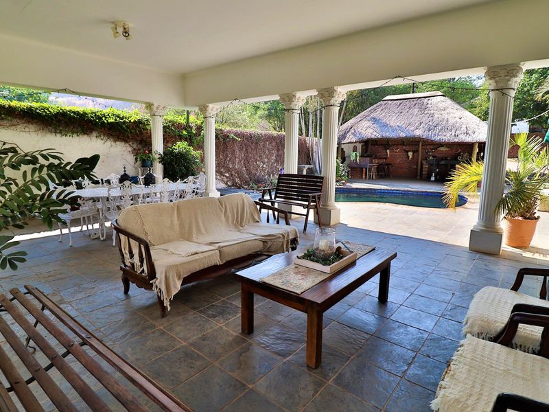 Spacious covered patio featuring stone tile flooring and decorative columns, overlooking a swimming pool and thatched lapa area.