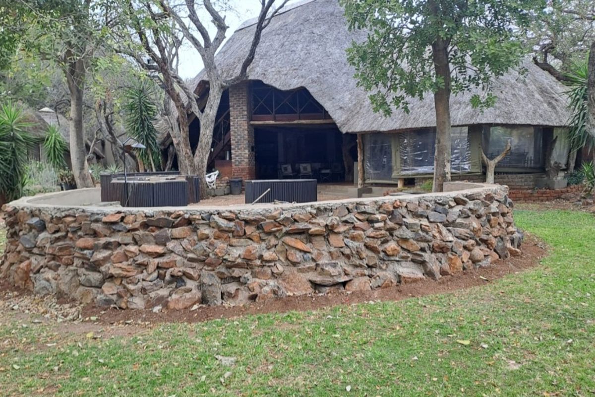 Rear view of a rustic farmhouse featuring a traditional thatched roof, a prominent stone retaining wall, and a covered patio area.