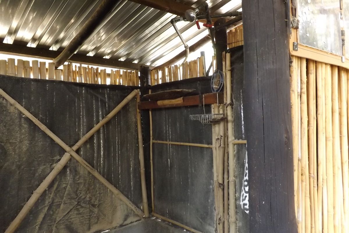 An outdoor-style rustic shower area featuring bamboo wall cladding, corrugated metal roofing, and exposed plumbing fixtures.