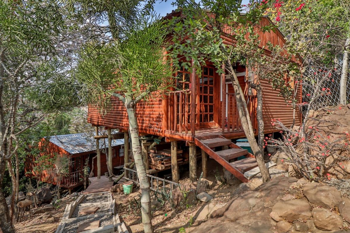 A rustic timber log cabin elevated on stilts, featuring a wooden deck, staircase access, and set within a natural, landscaped garden environment.