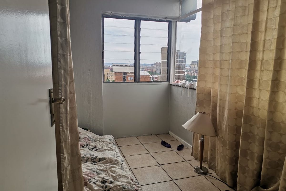A compact bedroom featuring tiled flooring and large windows with city views, offering a functional space with potential for modernization.