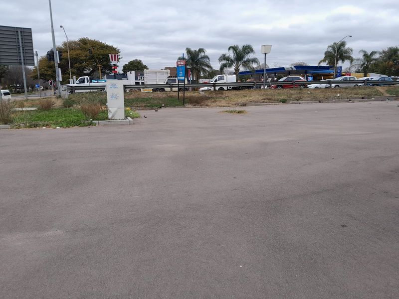 An open asphalt parking area with a guard rail and utility box, situated near a busy road and commercial infrastructure.