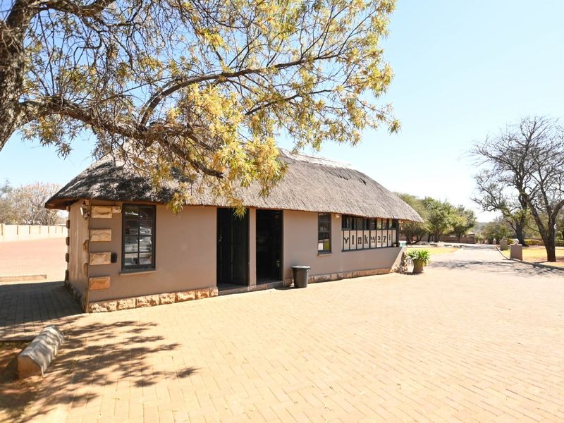A charming lodge building featuring traditional thatch roofing, stone accents, and a spacious paved entrance area, set within a natural landscape.