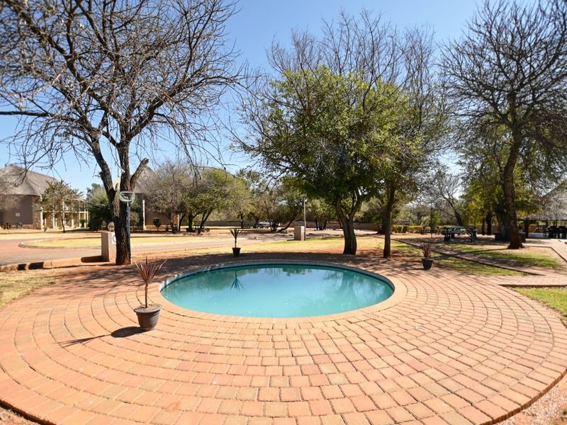 A circular swimming pool set within a landscaped garden area, featuring a decorative brick-paved surround and mature trees.