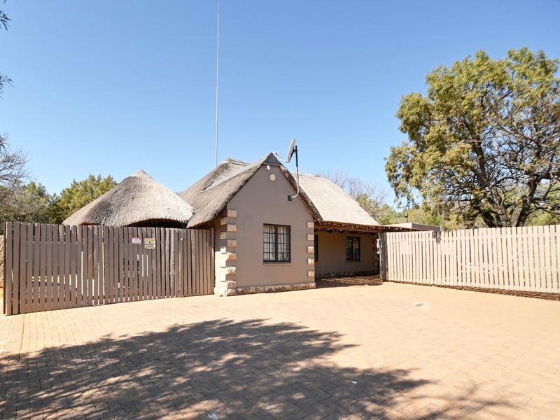 Front exterior view of a lodge featuring a traditional thatch roof, stone detailing, and a spacious paved driveway enclosed by timber fencing.