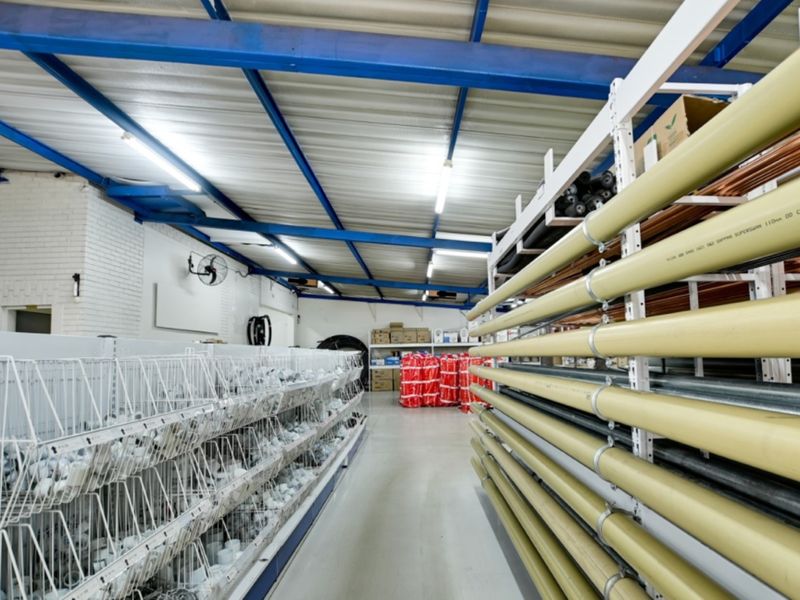 A well-maintained industrial retail utility space featuring exposed steel roof trusses, fluorescent lighting, and extensive fixed industrial shelving for inventory storage.