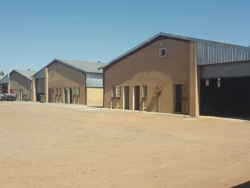 A row of industrial warehouse units featuring metal roofing, roller shutter doors, and a large unpaved yard area for vehicle access.