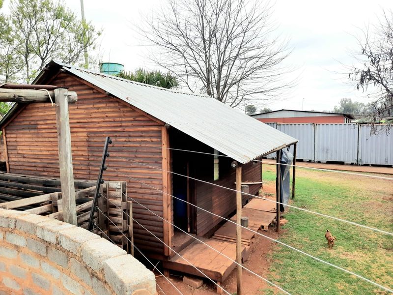 A timber-clad structure featuring a corrugated metal roof, a small wooden deck, and perimeter electric fencing within a garden setting.