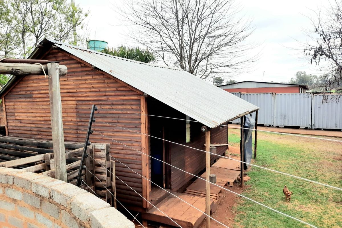 A timber-clad structure featuring a corrugated metal roof, a small wooden deck, and perimeter electric fencing within a garden setting.