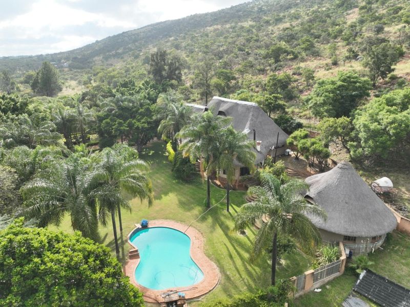 Aerial view of a farm property featuring a swimming pool, lush garden, and traditional thatch-roofed buildings set against a scenic hillside landscape.