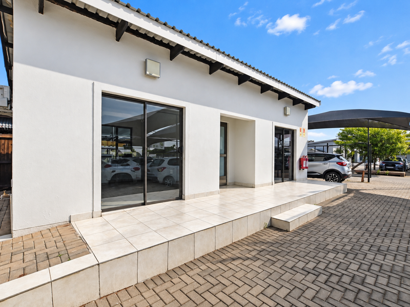 Exterior view of a modern office building featuring a tiled entrance patio, sliding glass doors, and paved parking area with shade cloth carports.