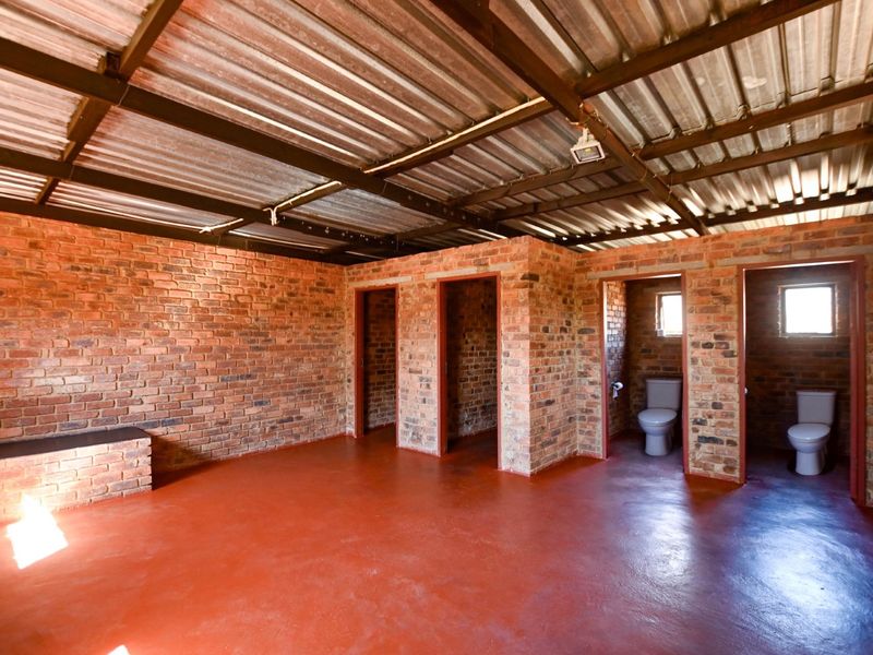 An open-plan utility or industrial space featuring exposed brick walls, a corrugated metal roof, and two private toilet cubicles with a wash basin.