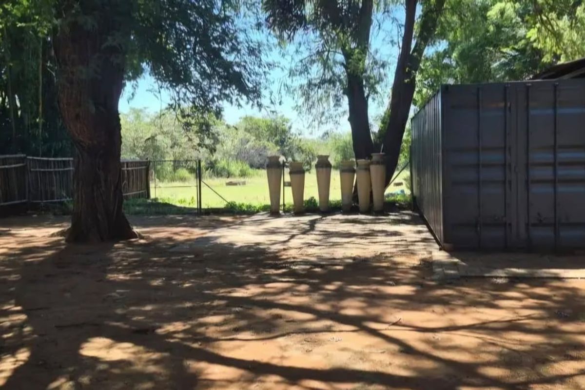 A rural outdoor area featuring a shipping container, a metal-roofed carport structure, and natural tree shading on a dirt and concrete surface.