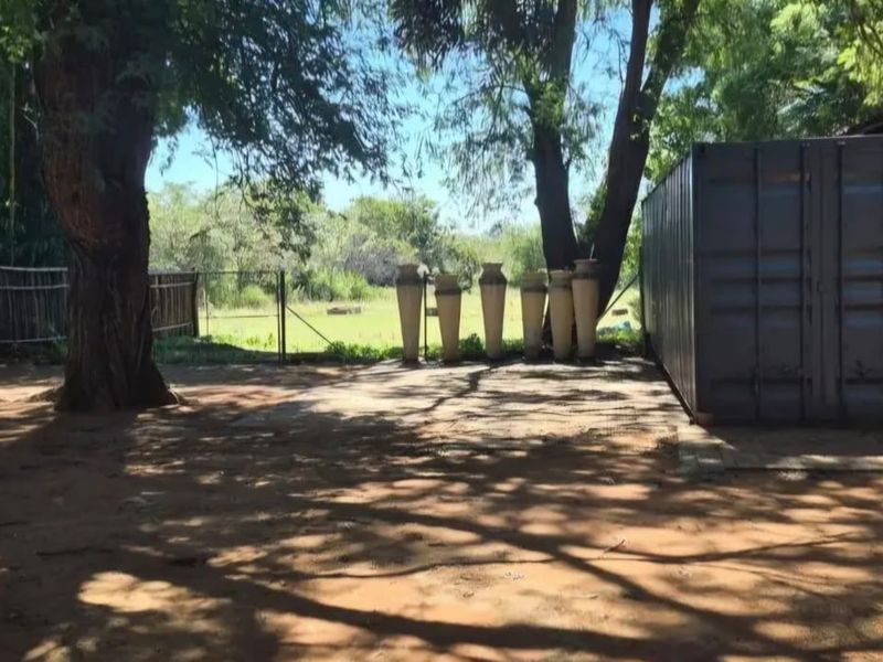 A rural outdoor area featuring a shipping container, a metal-roofed carport structure, and natural tree shading on a dirt and concrete surface.