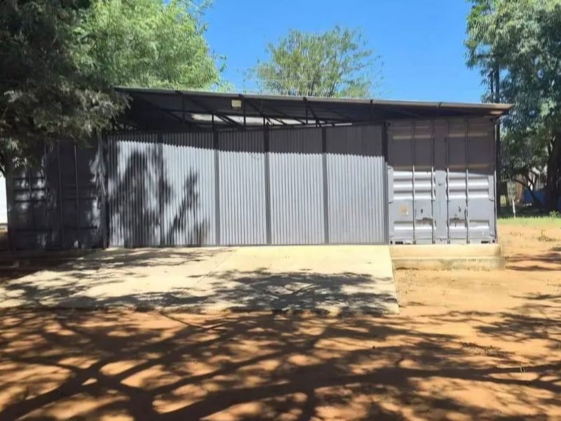 A functional garage structure constructed from shipping containers with corrugated metal cladding and a concrete apron, situated on a farm property.