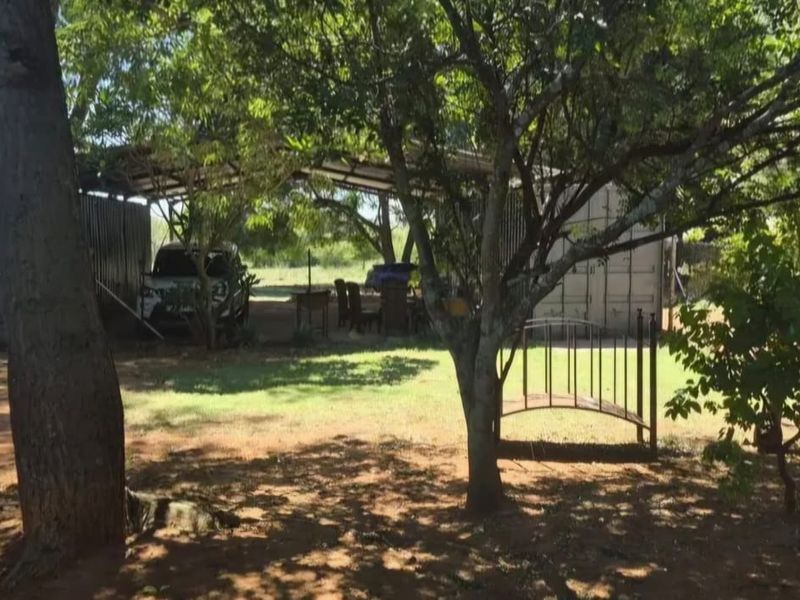 A shaded outdoor area featuring a metal carport structure, shipping containers, and a decorative garden bridge set within a natural farm landscape.
