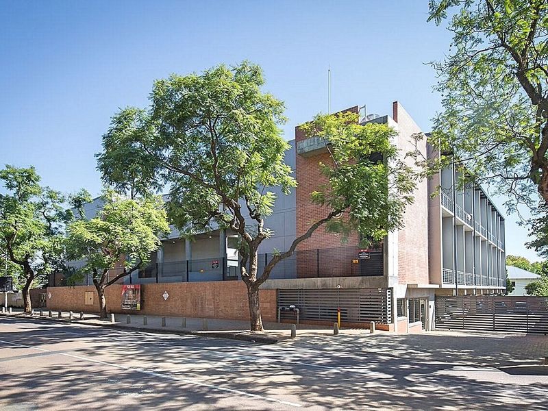 Street-level view of a multi-story apartment building featuring a brick facade, secure gated entrance, and paved driveway access.
