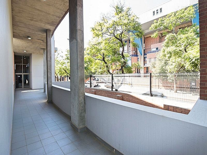 A spacious covered balcony featuring concrete structural elements, tiled flooring, and a view overlooking the street and surrounding complex.