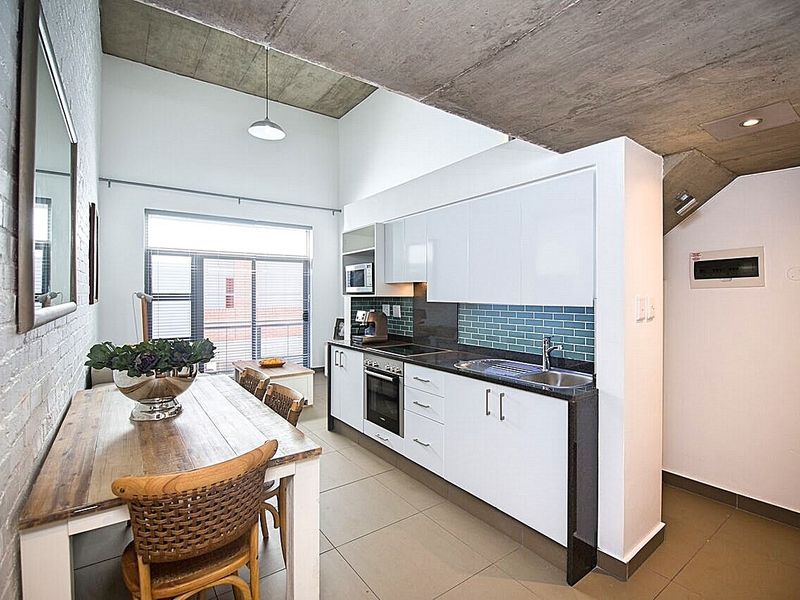 Modern open-plan kitchen and dining area featuring high exposed concrete ceilings, sleek white cabinetry, stone countertops, and a stylish tiled backsplash.
