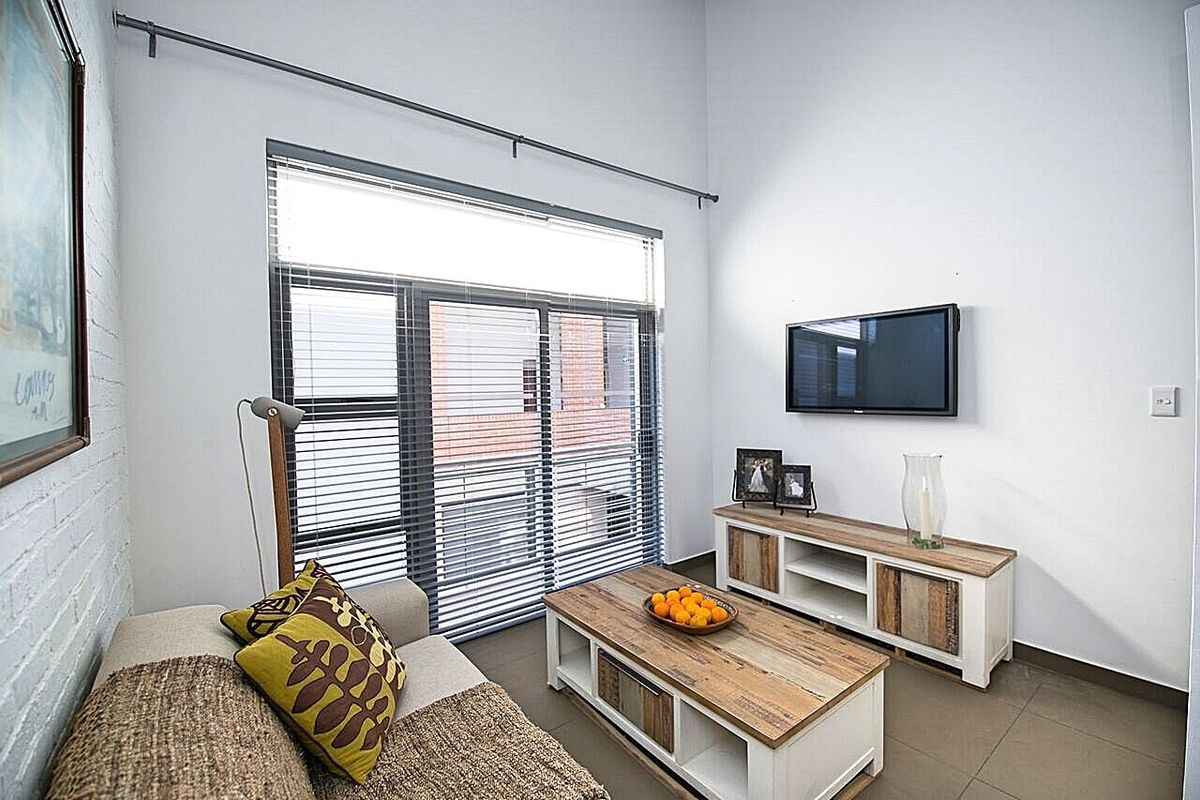 A bright living room featuring high ceilings, large windows with venetian blinds, and neutral tiled flooring.
