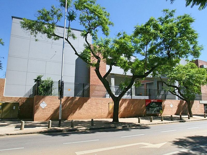 Street view of the apartment complex featuring a brick boundary wall, security fencing, and mature trees along the sidewalk.