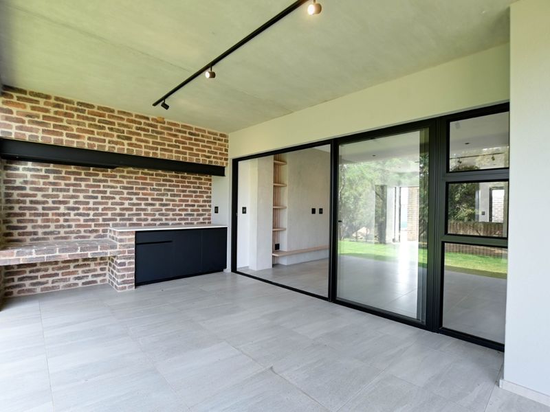 A modern covered patio featuring an exposed brick wall, built-in braai, track lighting, and large sliding glass doors leading to the interior living space.