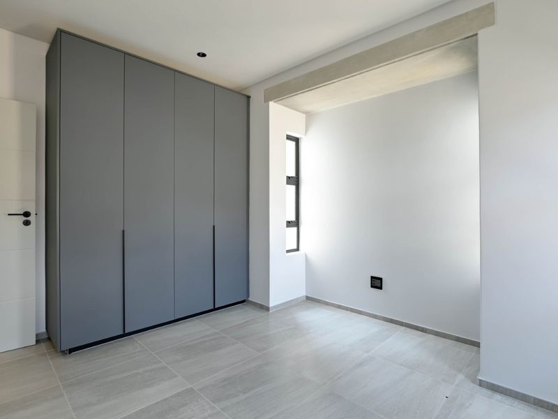 A modern, empty bedroom featuring sleek grey built-in wardrobes, neutral tiled flooring, and an exposed concrete architectural beam detail.