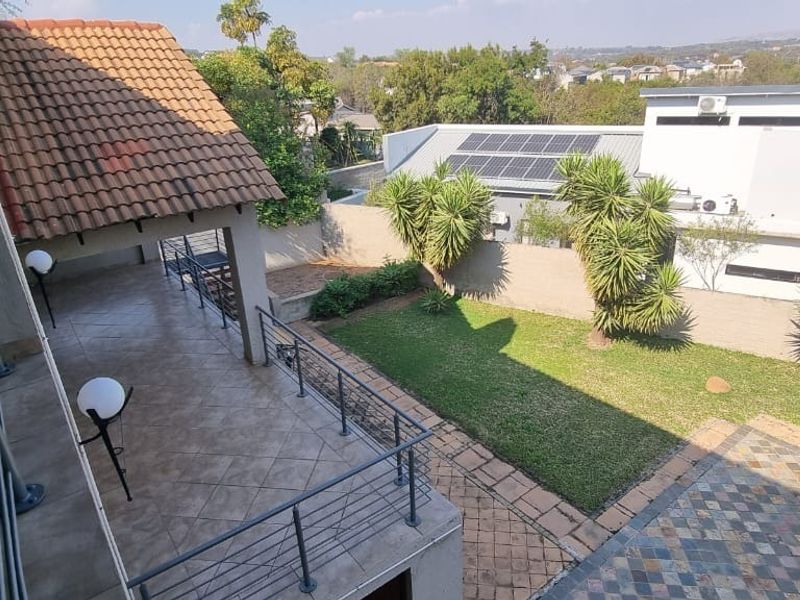 An elevated view of a rear garden featuring a covered tiled patio with metal balustrades, a lawn area, and paved pathways.