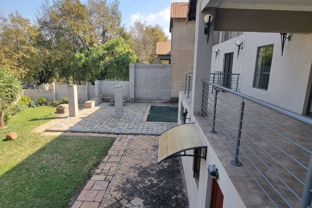 Rear exterior view featuring a covered balcony, paved patio area, swimming pool, and landscaped garden with brick boundary walls.