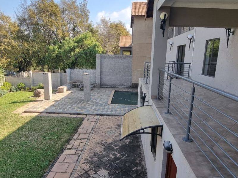 Rear exterior view featuring a covered balcony, paved patio area, swimming pool, and landscaped garden with brick boundary walls.