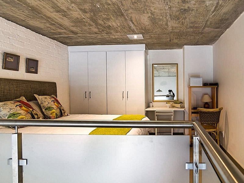 A modern bedroom featuring an exposed concrete ceiling, painted brick wall, and built-in wardrobe, overlooking a lower level via a glass and steel balustrade.