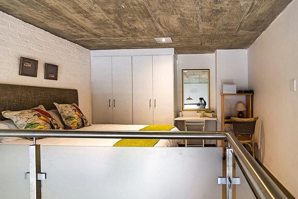 A modern bedroom featuring an exposed concrete ceiling, painted brick wall, and built-in wardrobe, overlooking a lower level via a glass and steel balustrade.