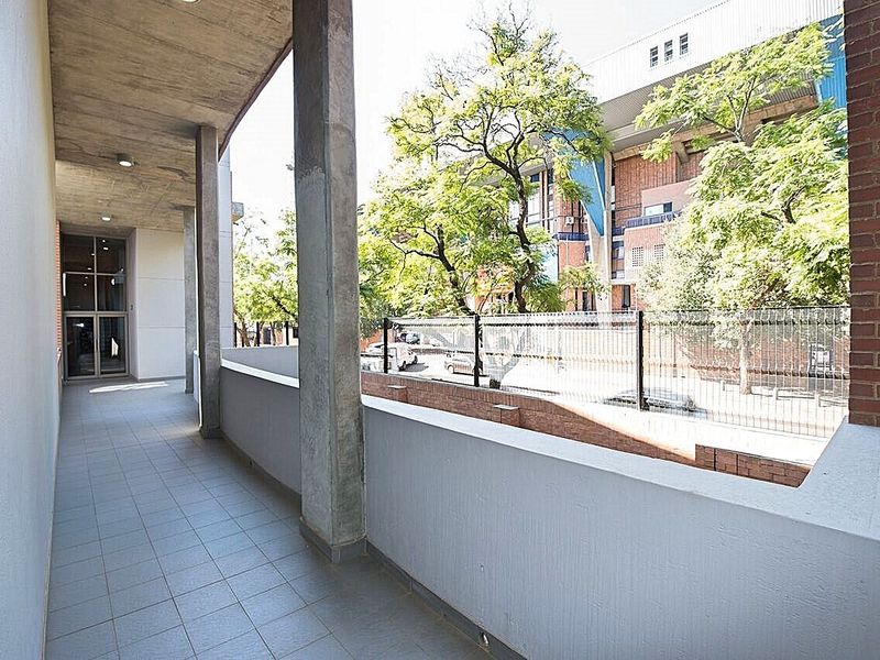 A covered balcony featuring concrete structural elements, tiled flooring, and a brick wall, overlooking a secure outdoor area with mature trees.