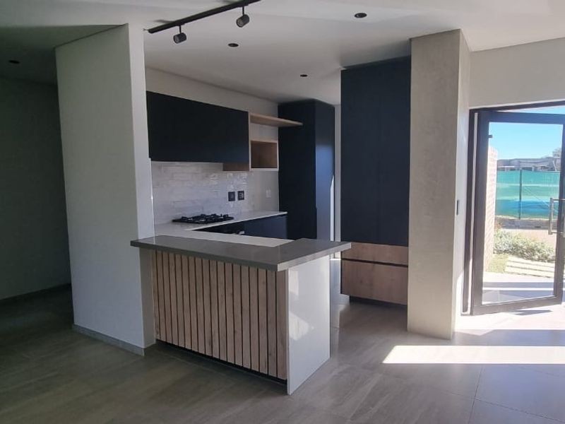 Modern open-plan kitchen featuring sleek dark cabinetry, stone benchtops, and a breakfast bar, seamlessly connecting to the outdoor area through a glass door.