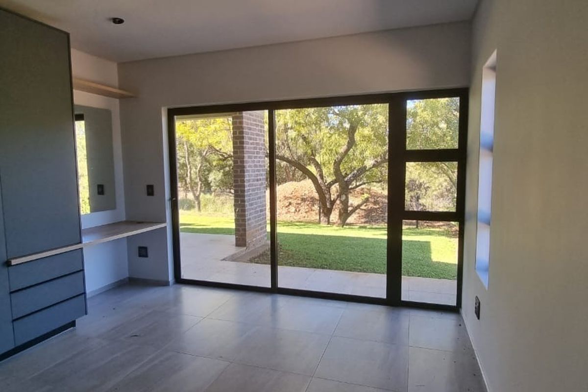 A bright, modern bedroom featuring built-in cupboards, a desk area, and large sliding glass doors that open onto a garden view.