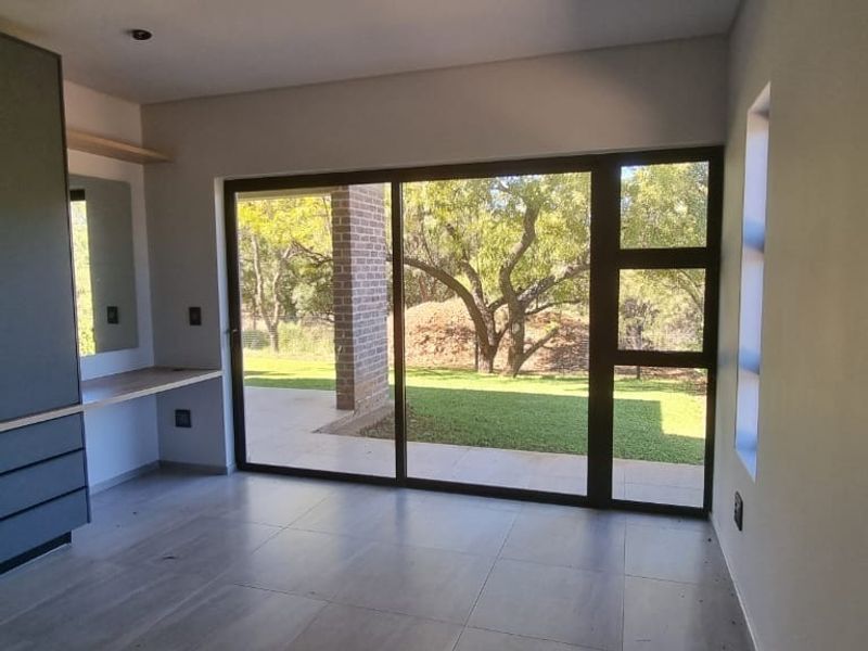 A bright, modern bedroom featuring built-in cupboards, a desk area, and large sliding glass doors that open onto a garden view.