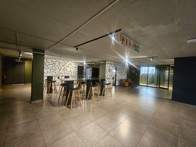 A modern communal clubhouse area featuring tiled flooring, exposed concrete ceilings, and industrial-style track lighting, suitable for social gatherings or building residents.