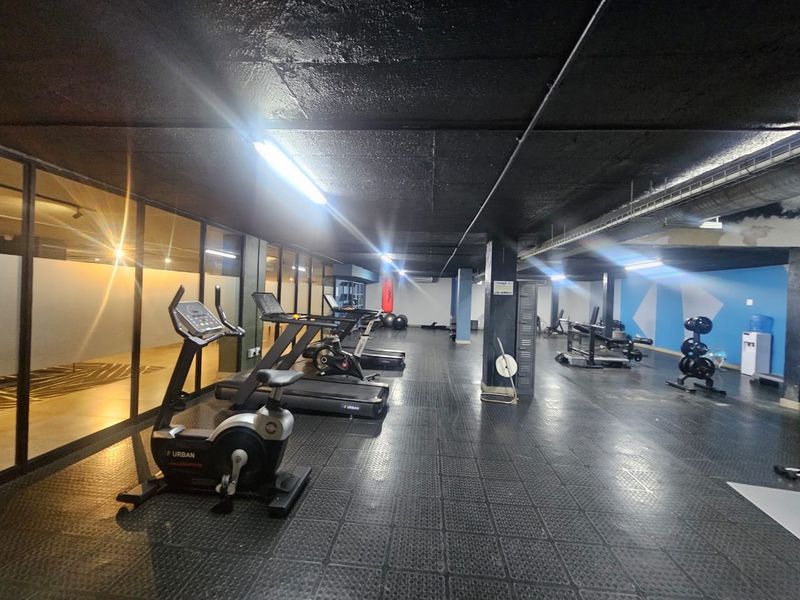A spacious apartment gym facility featuring rubber flooring, exposed industrial-style ceilings, and glass partitions, equipped with various fitness machines.