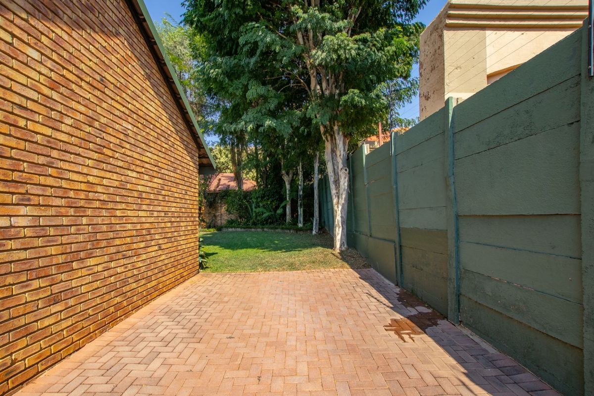 A side exterior view featuring a paved walkway alongside a brick building wall, leading to a small rear garden area with mature trees.