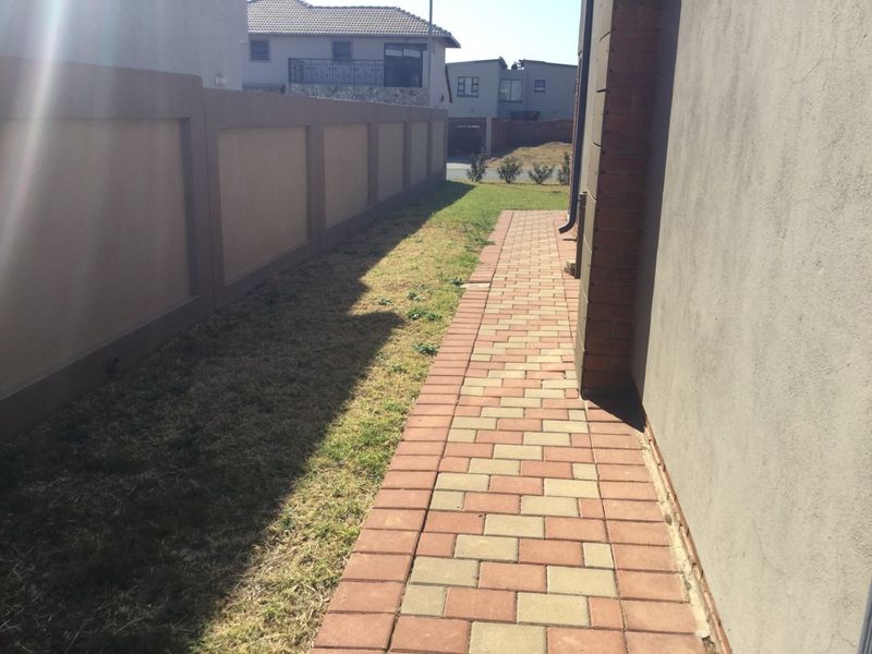 A side exterior view showing a paved walkway alongside the house, bordered by a boundary wall and a small lawn area.