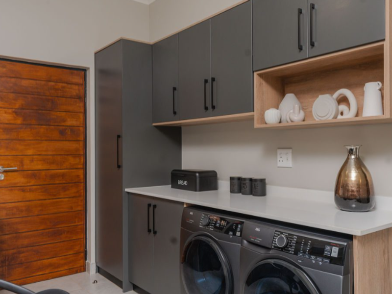 Modern laundry room featuring sleek dark grey cabinetry, timber-look open shelving, stone countertops, and a solid timber door.