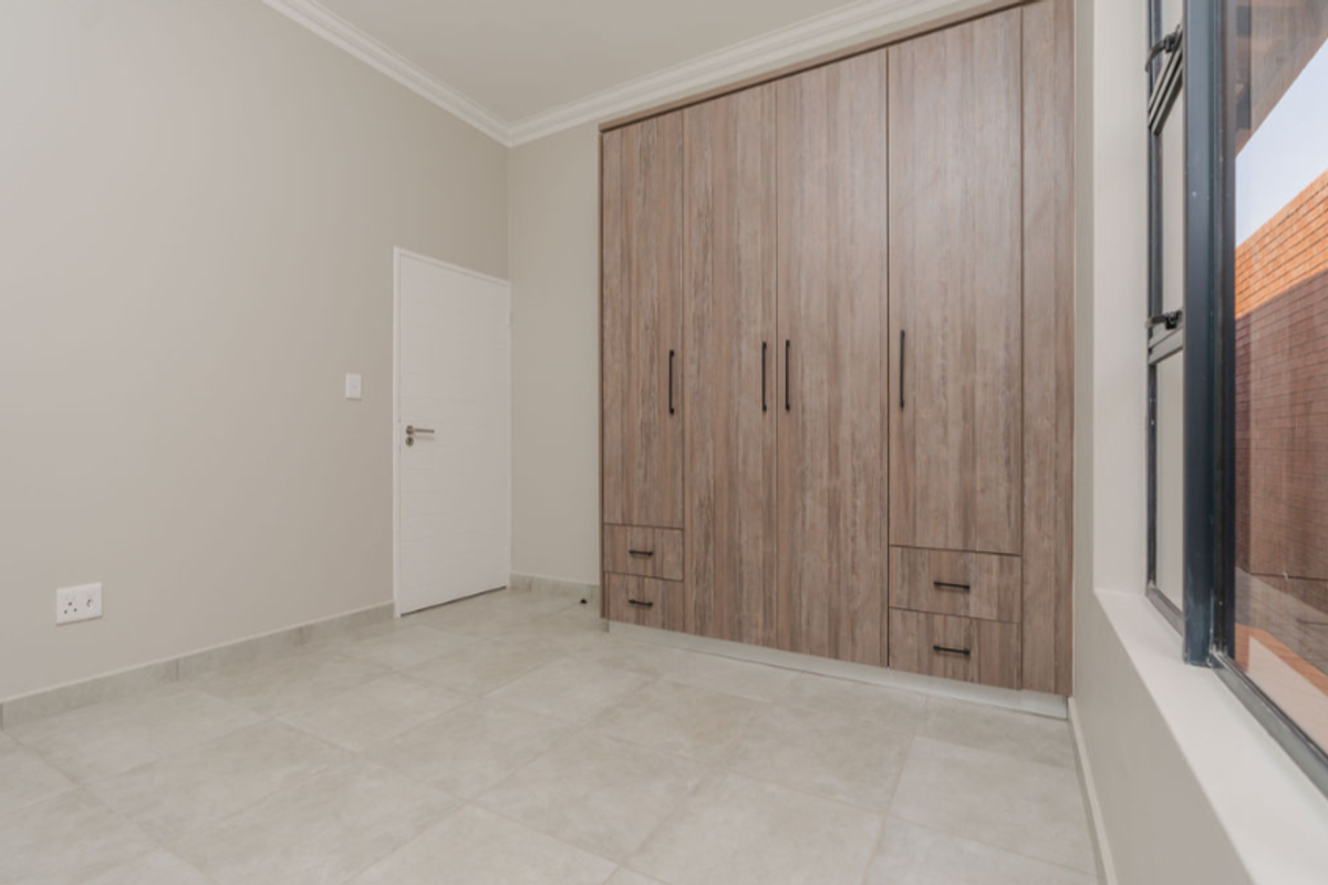 A bright, modern bedroom featuring neutral tiled flooring, built-in wooden cabinetry with integrated drawers, and large windows allowing for natural light.