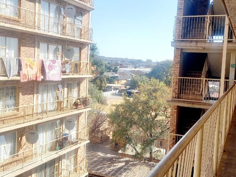 An exterior view of an apartment building balcony featuring tiled flooring and metal railings, overlooking the complex courtyard and adjacent building facade.