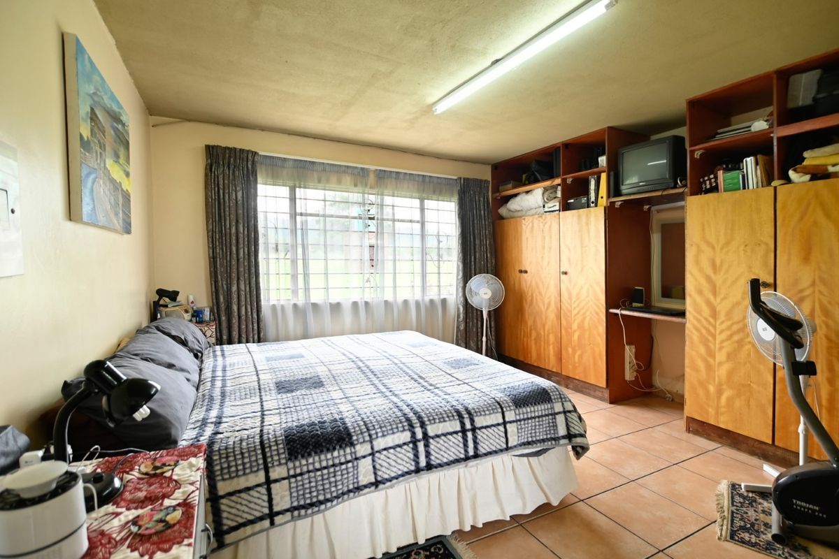 A bedroom featuring built-in wooden cupboards, tiled flooring, and a large window with security bars, offering a functional space with dated finishes.