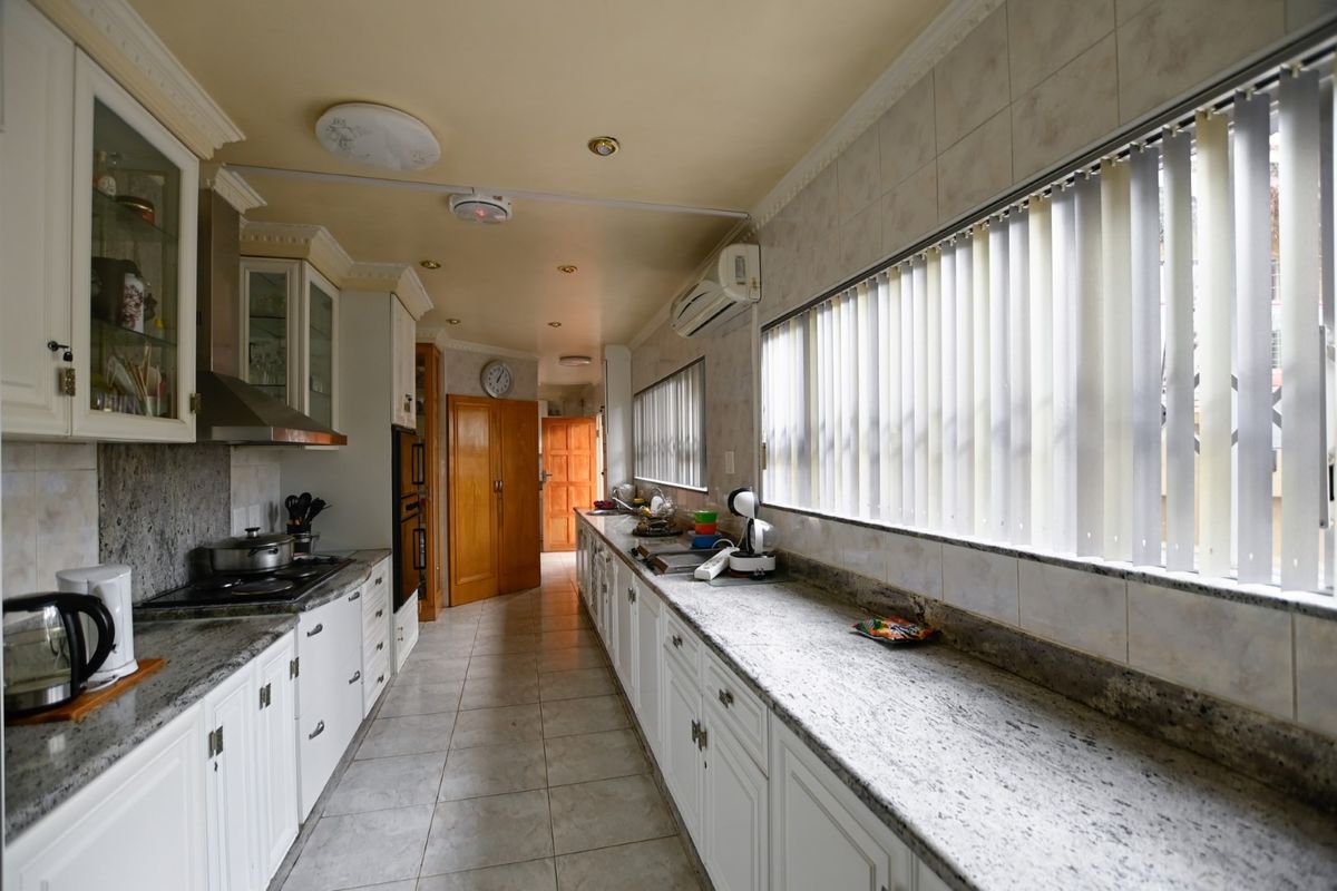 A long galley-style kitchen featuring granite countertops, extensive built-in cabinetry, a built-in oven, and a hob, with tiled flooring and vertical window blinds.