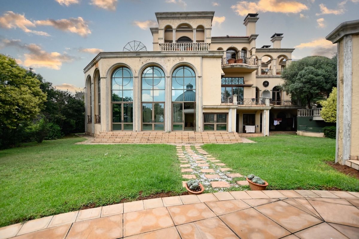 Rear view of a grand multi-story residence featuring large arched windows, multiple balconies, stone patio, and a spacious garden.