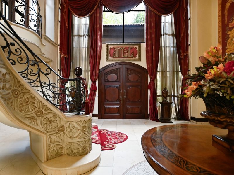 Grand entrance foyer featuring a sweeping staircase with ornate wrought iron railings, marble flooring, and impressive double-height windows with elegant drapery.