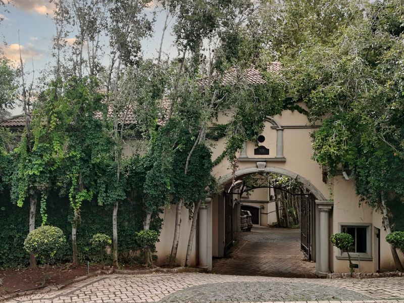 A grand arched stone entrance gateway with a paved driveway, featuring mature landscaping and a classic stucco facade.