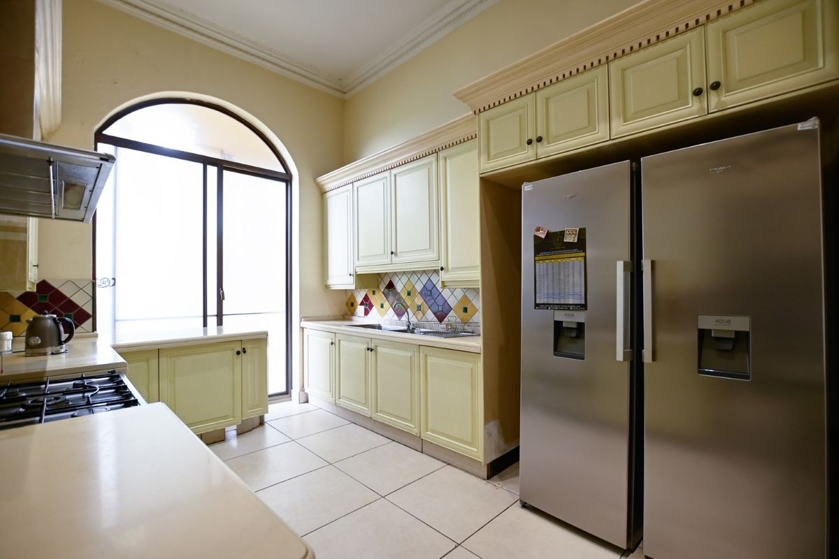 A functional kitchen featuring light-colored cabinetry, a decorative tiled backsplash, tiled flooring, and a large arched window providing natural light.
